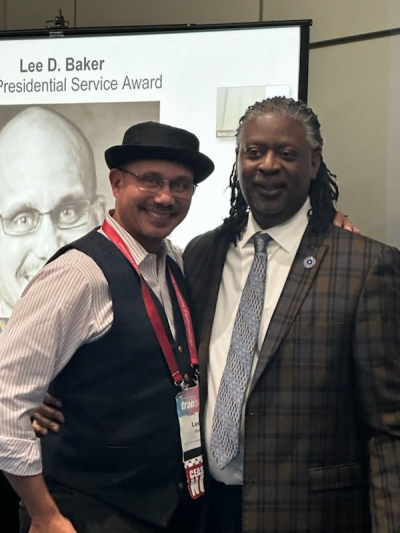 Two men smiling for the camera with screen behind them displaying photo of Lee Baker and title Lee D. Baker Presidential Service Award